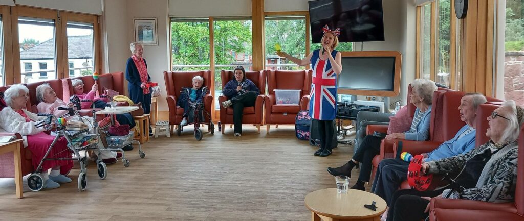 At Lear House, a group of elderly people sit in a lounge as Thelma, wearing a Union Jack outfit and hat, entertains them with a microphone and yellow gloves during a lively birthday celebration.