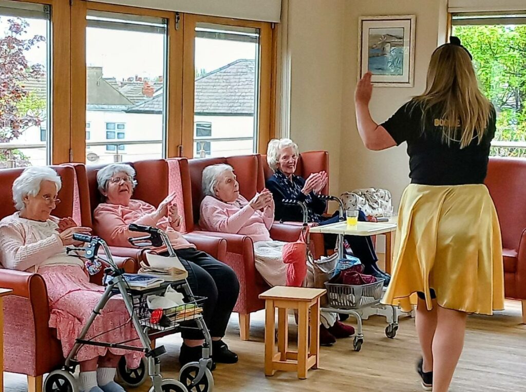A group of elderly women sit in armchairs at Lear House, watching a woman in a black top and gold skirt perform a lively Baby Boogie dance.