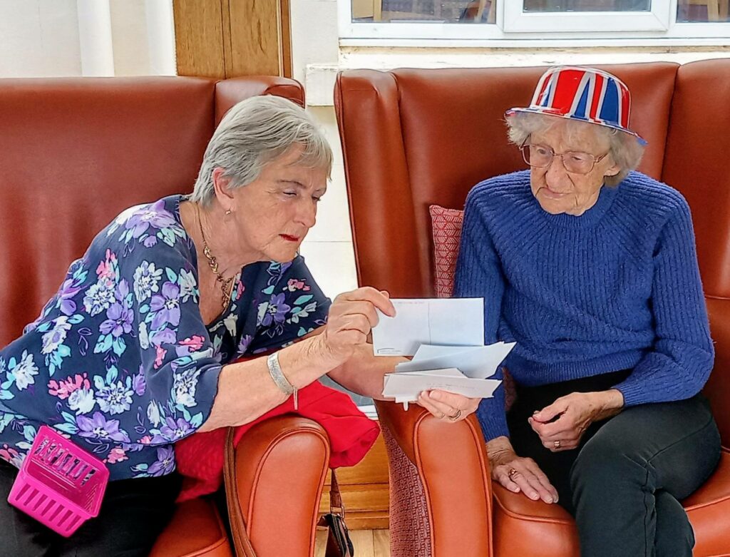 Two elderly women sit in armchairs; one is showing the other a stack of letters. The woman on the right, at Lear House, wears a Union Jack hat and blue jumper—showing true Best of British spirit.
