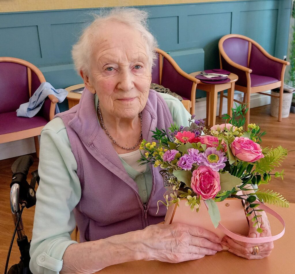Elderly woman with short white hair sits at a table, displaying her creativity as she holds a colourful bouquet of flowers at Lear House, with empty chairs and a walking frame in the background.
