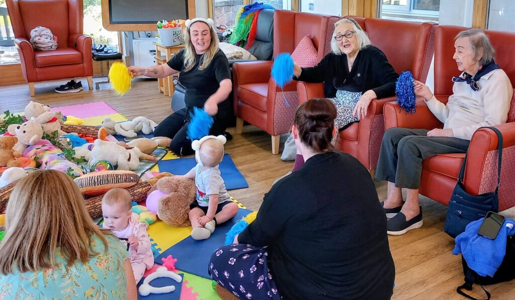 Older adults and babies engage on a colourful play mat with toys, while women seated nearby hold pom-poms, in a bright, communal room.