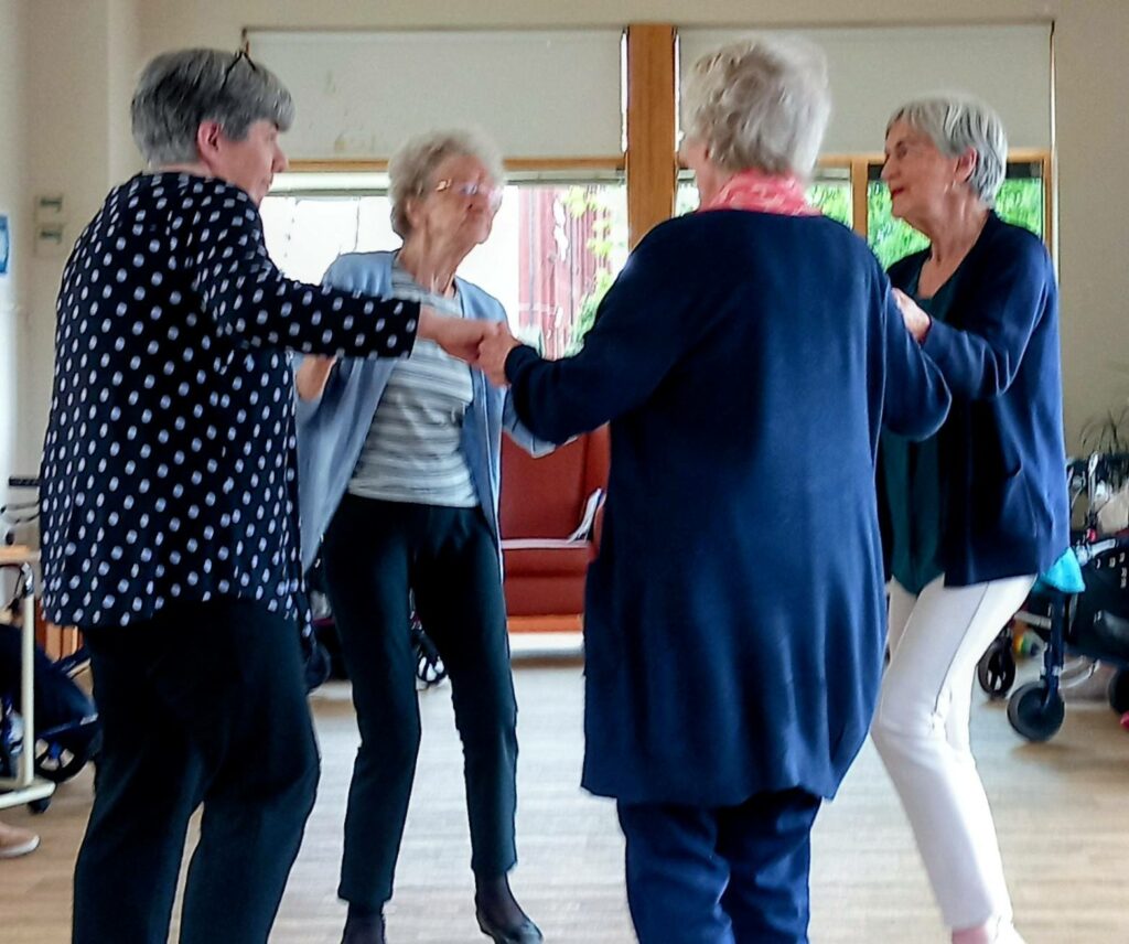 Four older women stand in a circle, holding hands and dancing together indoors at Lear House, natural light pouring through large windows—perhaps celebrating Thelma’s special birthday.