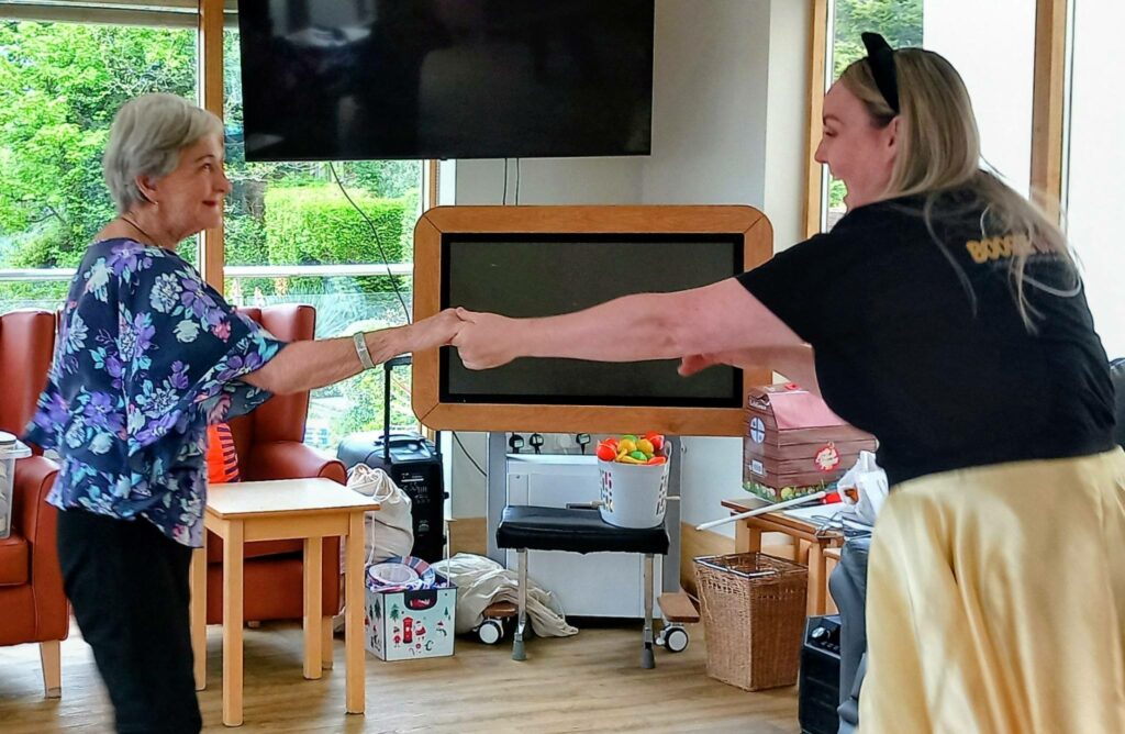Two women are holding hands and dancing in a light-filled room at Lear House, with chairs, a telly, and assorted items in the background—capturing the lively spirit of Baby Boogie and Best of British fun.