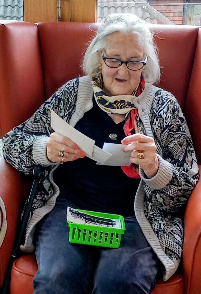 An elderly woman wearing glasses and a scarf sits in a red chair at Lear House, looking at several papers. A green plastic basket rests on her lap.