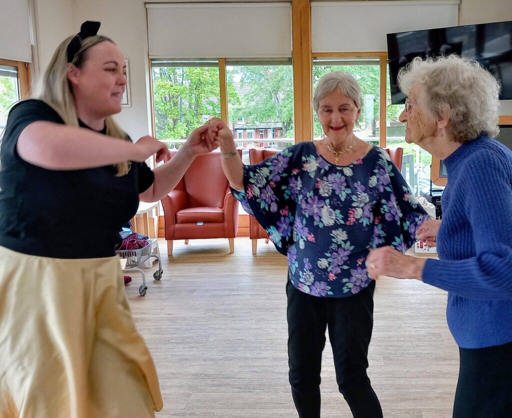Three women—one younger, two older—hold hands and dance together in a bright room with large windows and wooden floors at Lear House’s Baby Boogie, celebrating the Best of British.