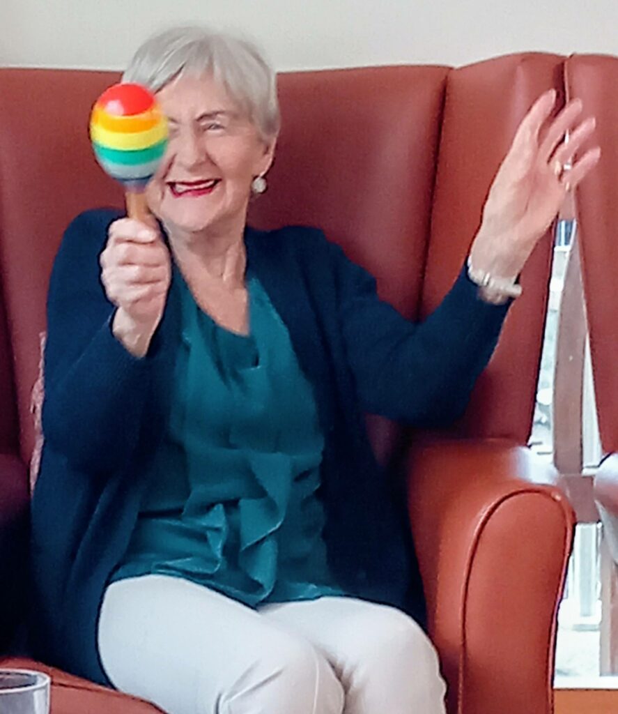An elderly woman named Thelma sits in an armchair at Lear House, smiling and raising her hand as she holds a colourful maraca during a joyful birthday celebration.