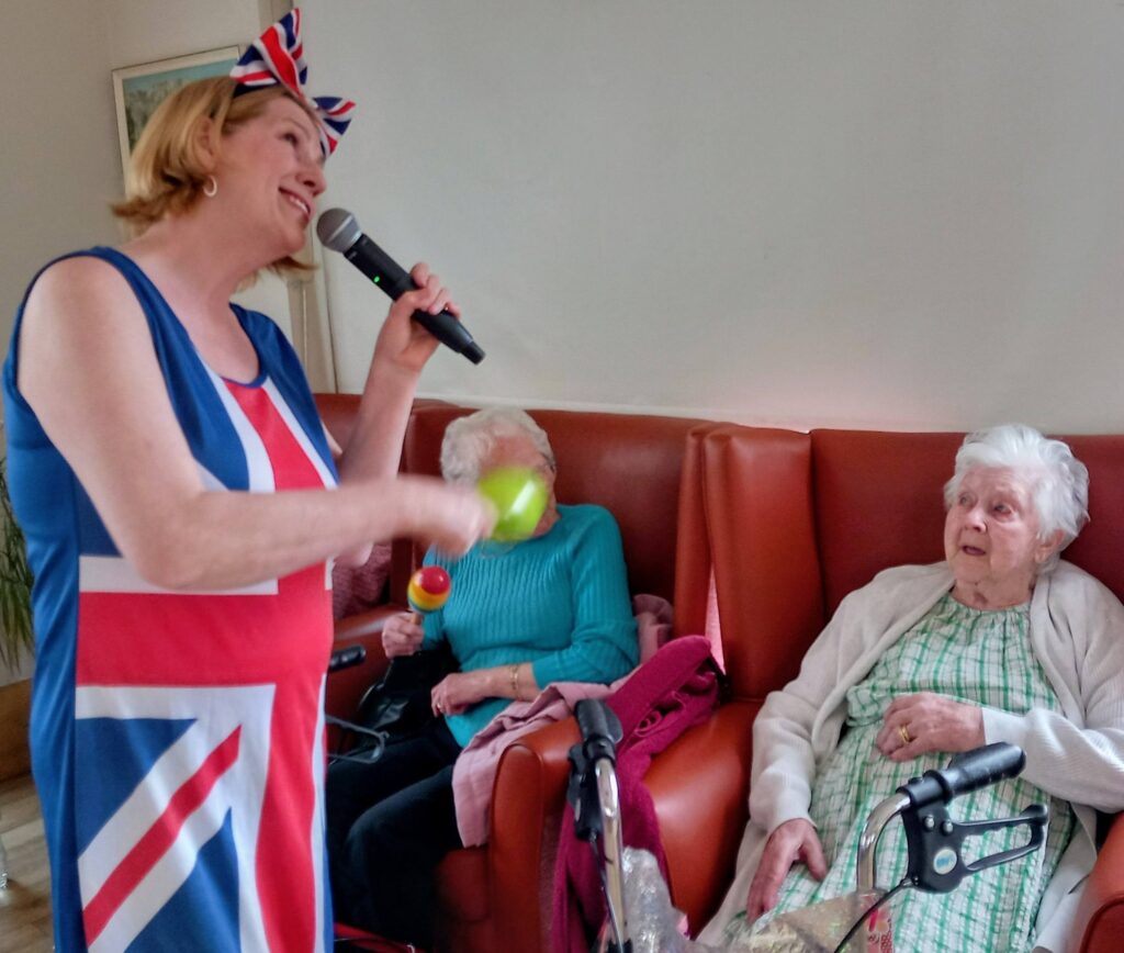 At Lear House, a woman in a Union Jack dress sings into a microphone and shakes a maraca for two elderly women seated on a sofa, one holding a maraca, during Thelma’s Birthday Celebration.