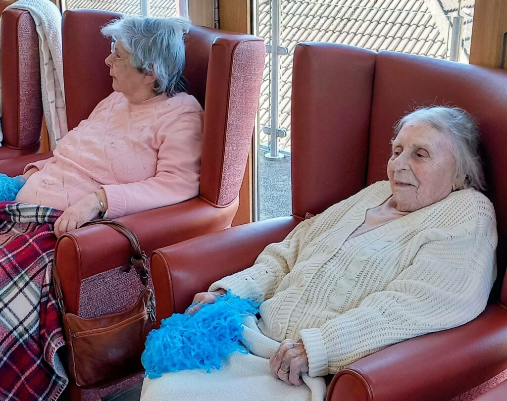 Two elderly women sit in red armchairs, each holding a blue pompom, with blankets over their laps and a window behind them offering a view of a tiled roof.