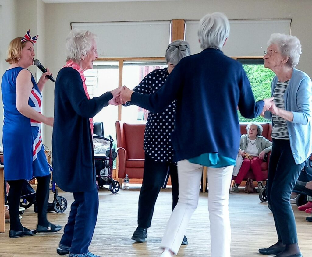 At Lear House, five elderly women dance in a circle indoors while Thelma, wearing a Union Flag dress, sings into a microphone during a joyful birthday celebration. Others are seated in the background.