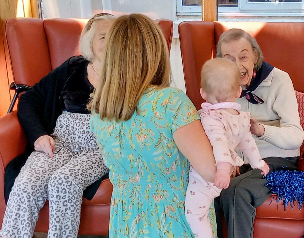 A woman holding a baby stands in front of two elderly women seated in red armchairs, engaging in conversation.