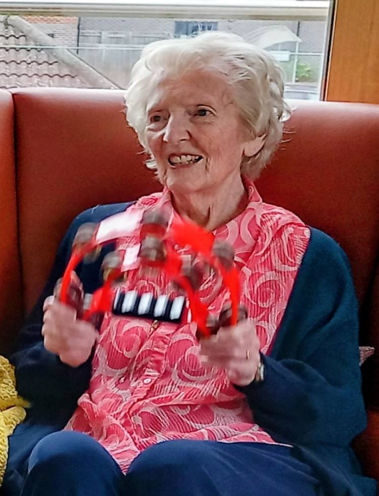 Thelma, an elderly woman, sits on a sofa at Lear House, smiling warmly and holding a red tambourine during a joyful birthday celebration.