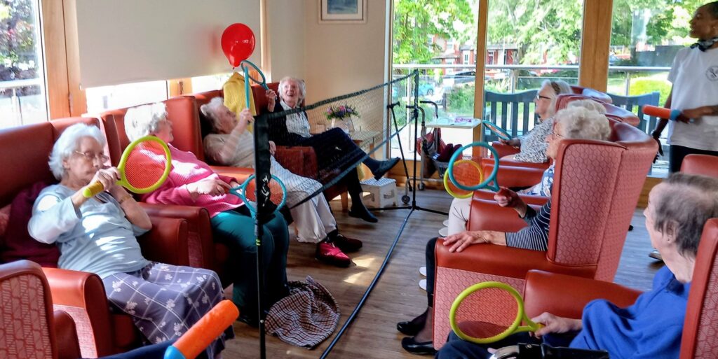 A group of elderly women seated in armchairs enjoy a lively game of Balloon Tennis with racquets and a net indoors, while others watch and a staff member assists in the background, promoting fun and flexibility.