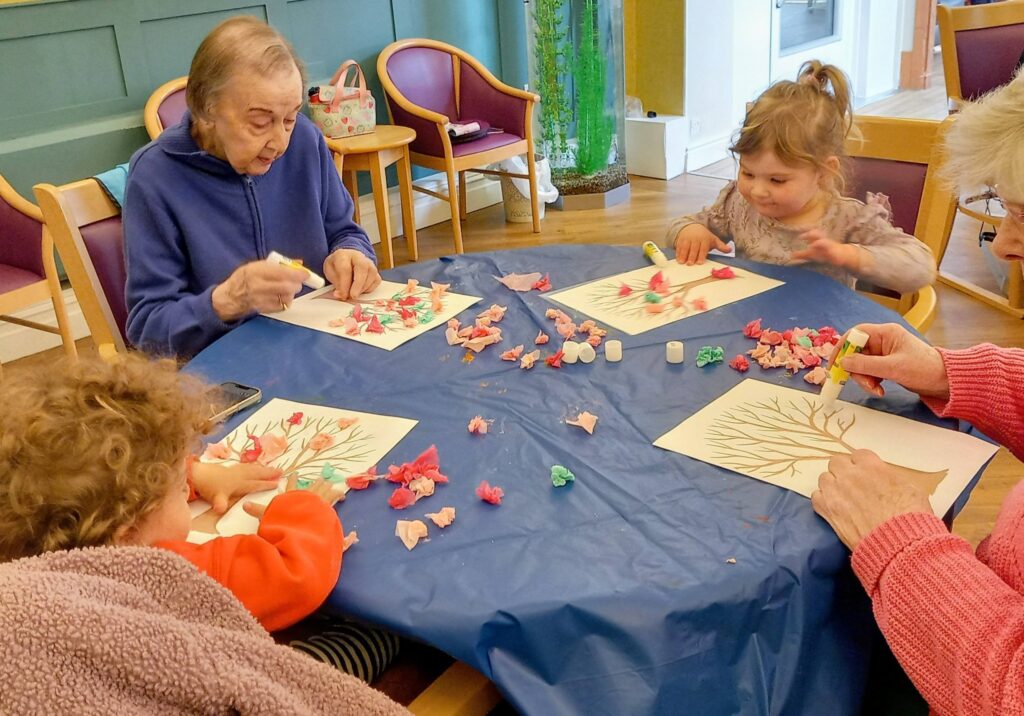 Two older adults and two young visitors sit at a table creating tree-themed crafts with paper and glue, enjoying a colourful morning together in a communal room.