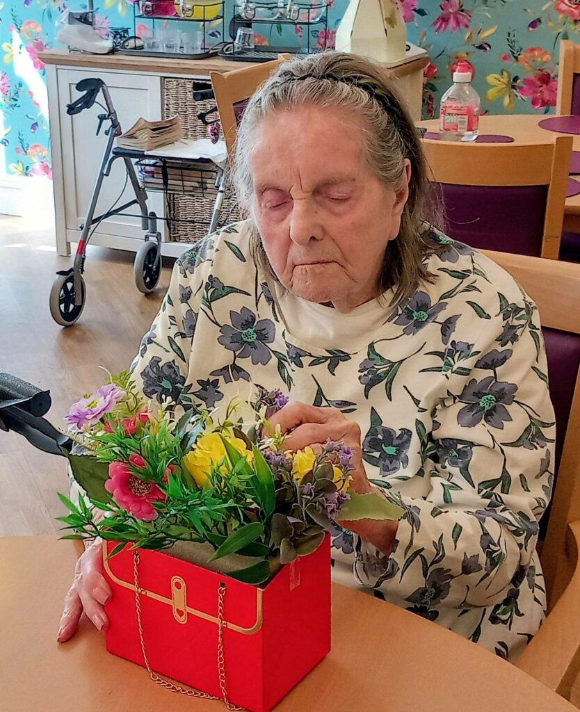 An elderly woman with grey hair shows creativity as she arranges artificial flowers in a red handbag-shaped container at Lear House. A walking frame and floral wallpaper can be seen in the background.