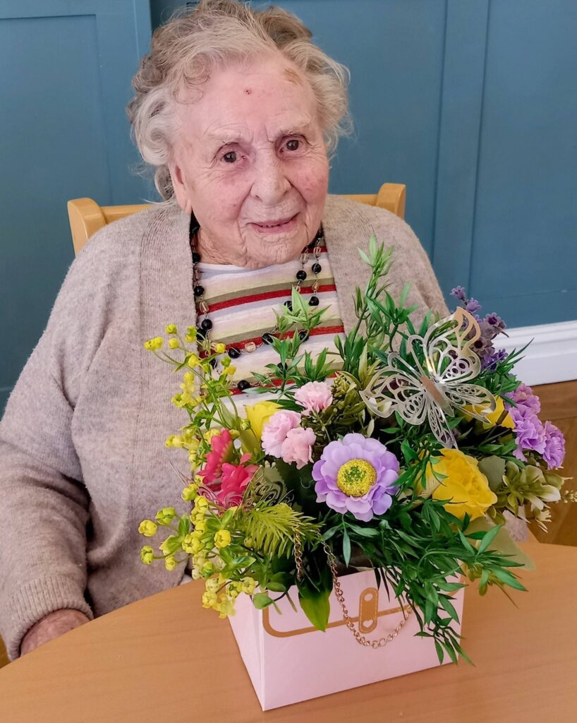 Elderly woman seated at a table, smiling, displaying her creativity with a colourful bouquet of artificial flowers arranged in a pink box at Lear House.