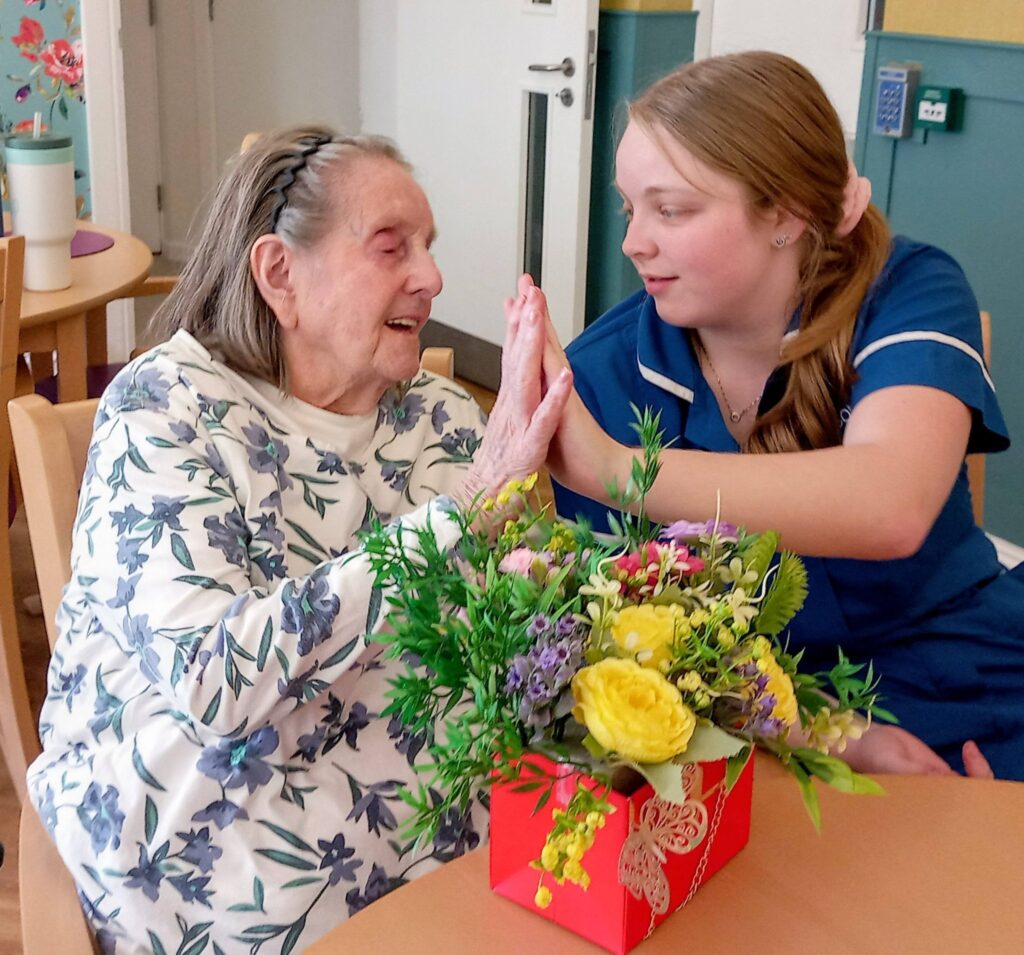An elderly woman and a young nurse sit at a table with a flower arrangement at Lear House, holding hands and smiling warmly at each other, sharing a creative and heartfelt moment.