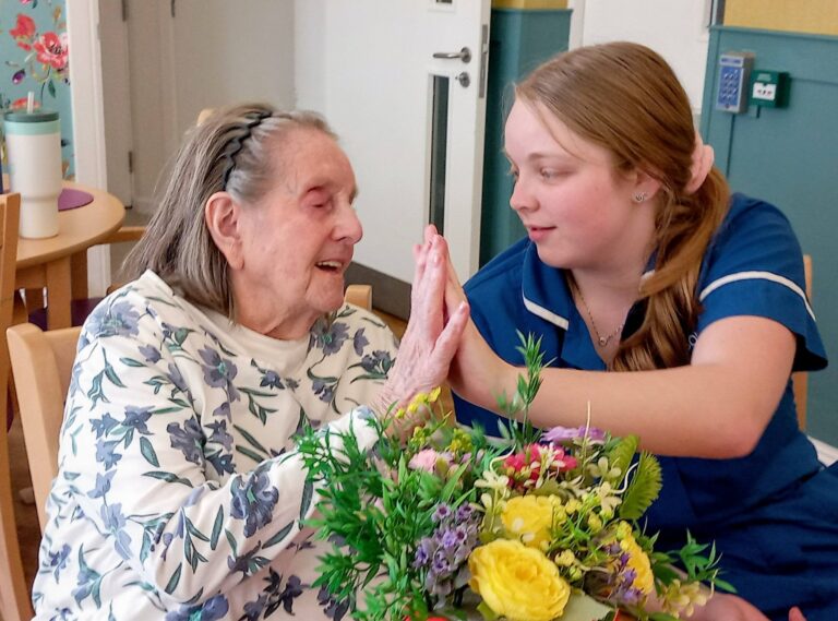 An elderly woman and a young nurse sit at a table with a flower arrangement at Lear House, holding hands and smiling warmly at each other, sharing a creative and heartfelt moment.