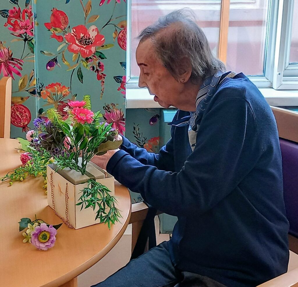 An elderly person sits at a table in Lear House, creatively arranging artificial flowers in a box. Floral wallpaper and a window can be seen in the background.