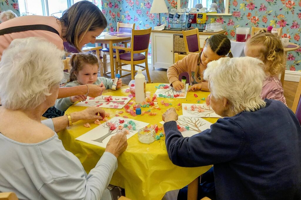 Children, young visitors, and elderly women sit around a yellow table making arts and crafts with paper, glue, and felt-tip pens during a colourful morning in a vibrant, flower-themed room.
