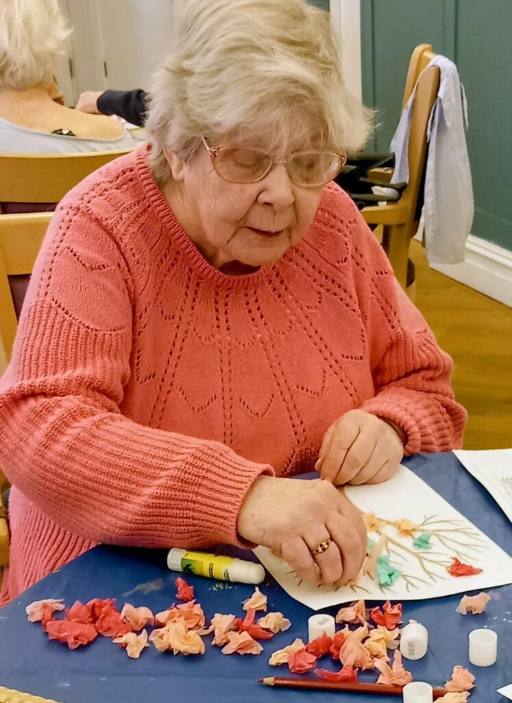 An elderly woman wearing glasses and a pink jumper enjoys a wonderful, colourful morning creating a craft project with coloured tissue paper at a table.