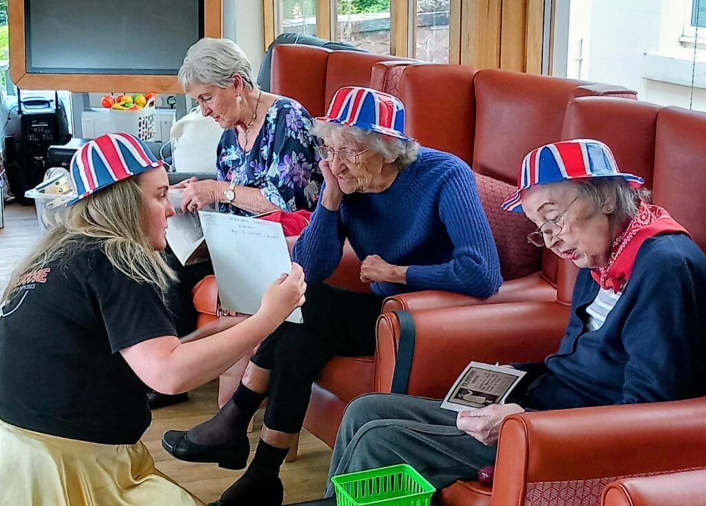 A woman at Lear House shows a sheet of paper to three elderly people wearing Union Jack hats on red chairs, celebrating the Best of British, whilst another woman writes in the background.