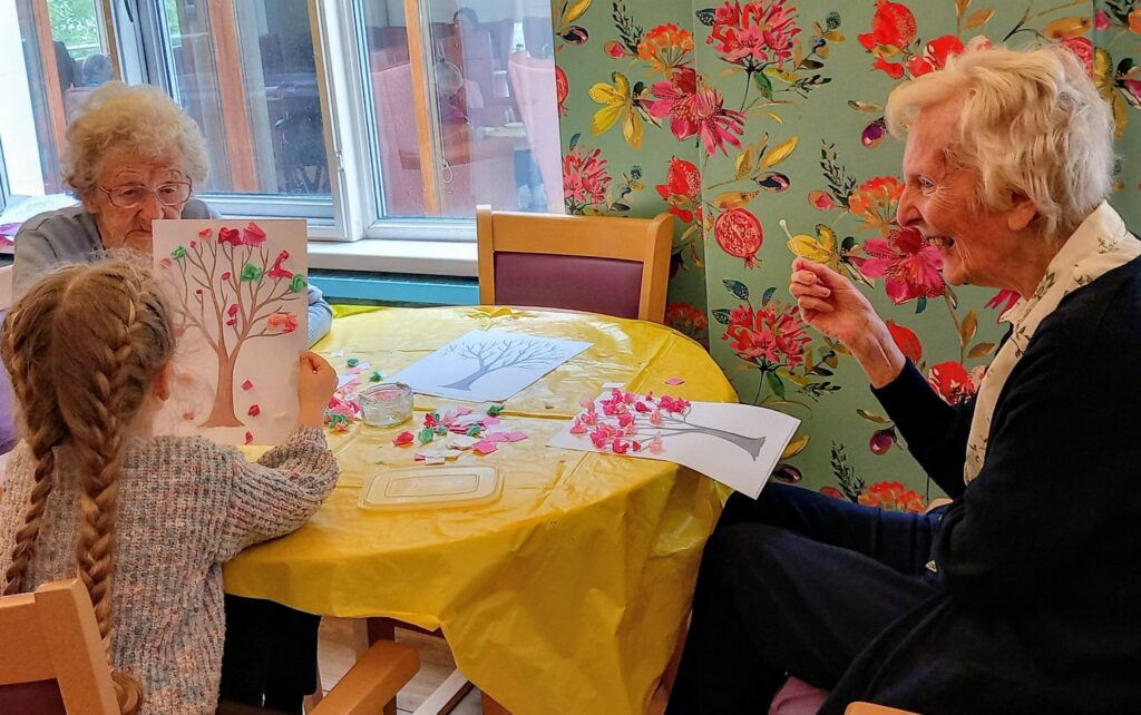 Two elderly women and a young girl, visitors to the community centre, sit at a table covered with art supplies, making tree-themed crafts on a colourful morning in a room decorated with floral wallpaper.