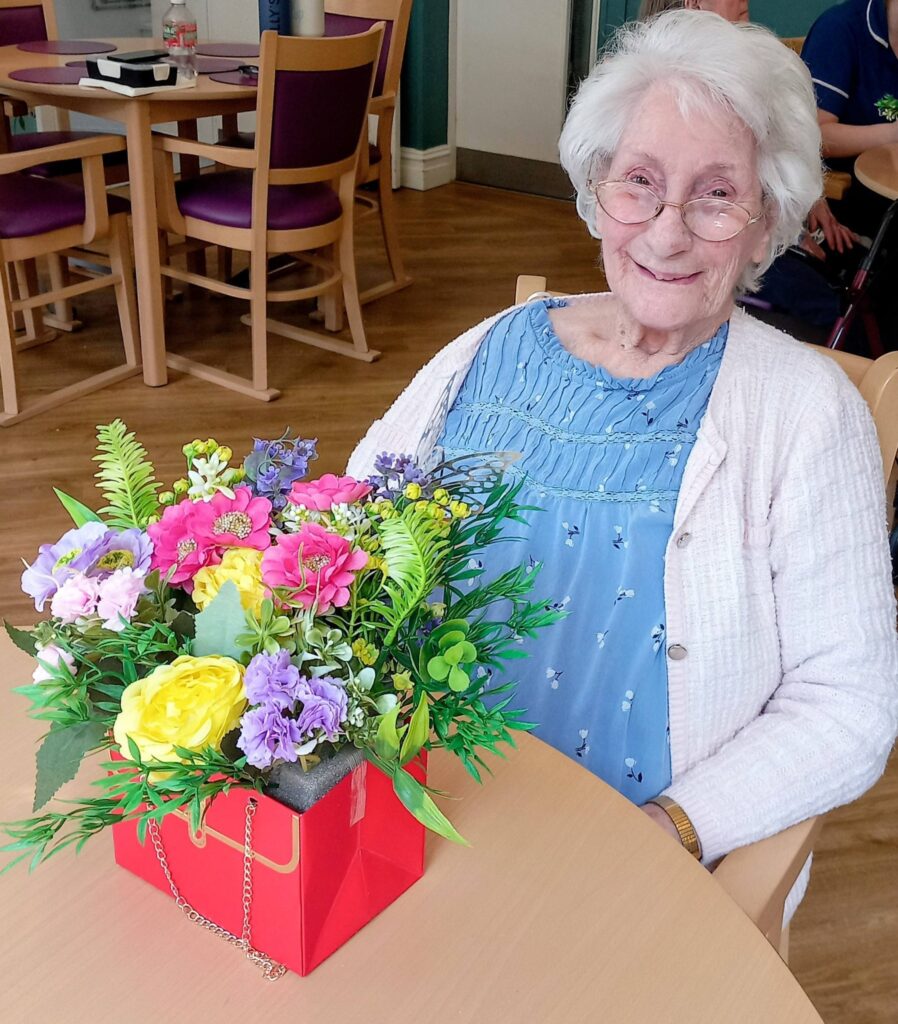 Elderly woman wearing glasses sits at a table, smiling, with a colourful bouquet of artificial flowers arranged in a red box before her, highlighting creativity at Lear House.