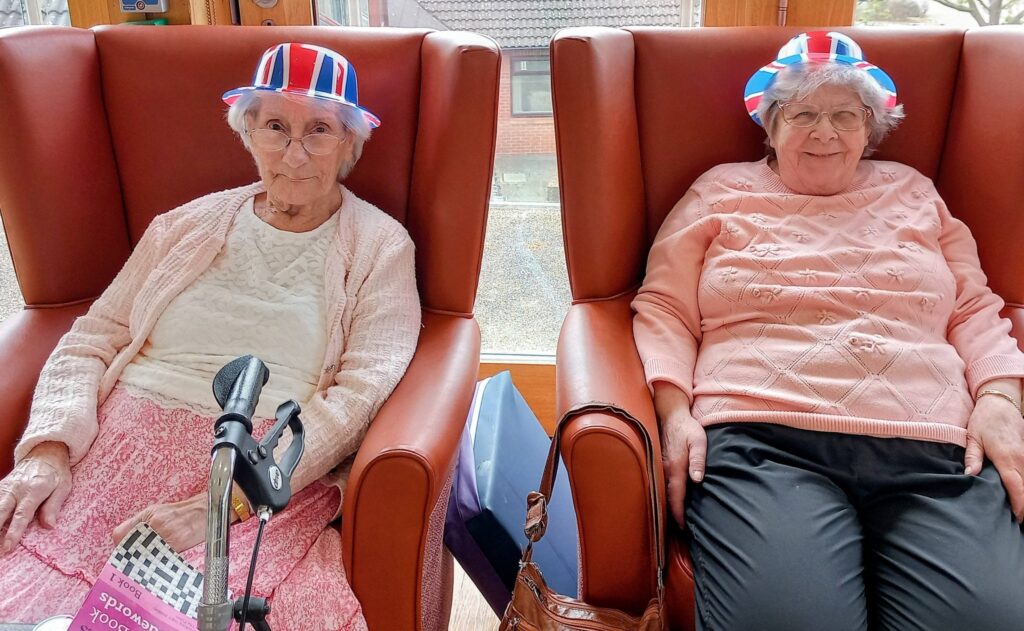 Two elderly women sit in armchairs at Lear House, sporting red, white, and blue hats. One holds a walking stick and a book; both wear light pink jumpers and spectacles, celebrating the Best of British spirit.
