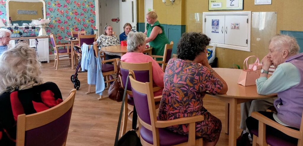 A group of elderly women sit chatting around tables in the brightly lit common room at Lear House, where creativity flourishes amidst colourful wallpaper and cheerful noticeboards.