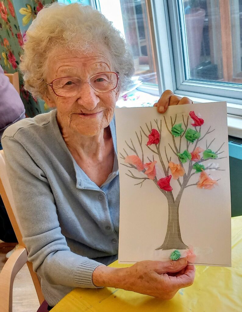 An elderly woman wearing glasses smiles on a colourful morning, holding a handmade artwork of a tree adorned with splendid, vibrant tissue paper leaves.