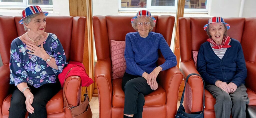 Three elderly women sit in high-backed chairs at Lear House, each wearing red, white, and blue hats, smiling together indoors with windows behind—a proper Best of British moment.
