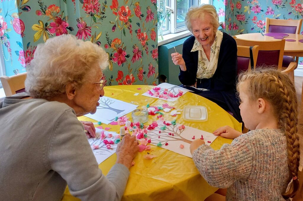 Two elderly women and a young girl enjoy a colourful morning crafting paper and tissue flowers at the table. Beautiful floral wallpaper adds brightness to the scene behind these young visitors.