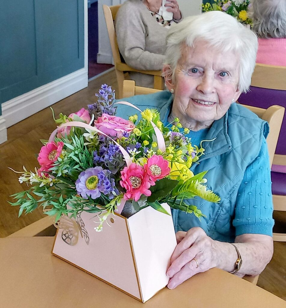 Elderly woman seated at a table in Lear House, smiling and holding a colourful flower arrangement in a box, demonstrating her creativity.