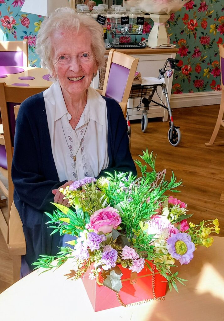 An elderly woman sits at a table in Lear House, surrounded by a colourful floral arrangement and floral wallpaper—an inspiring space that celebrates creativity and comfort with wooden furniture.