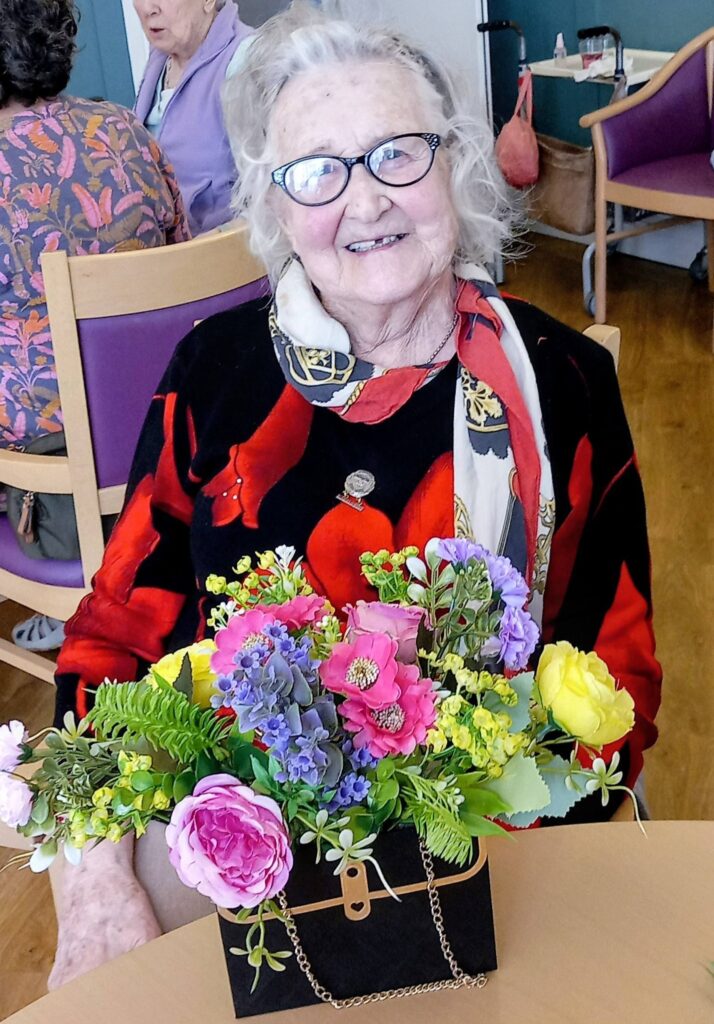 Elderly woman wearing glasses and a colourful scarf smiles whilst seated at a table in Lear House, her creativity evident in the vibrant floral arrangement displayed before her.