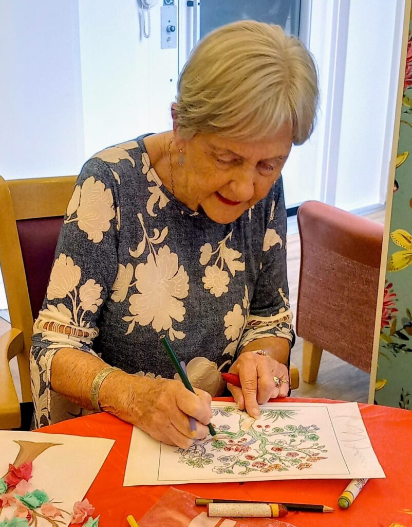 An elderly woman enjoys a wonderful, colourful morning as she sits at a table, colouring a floral design on paper with coloured pencils and markers.