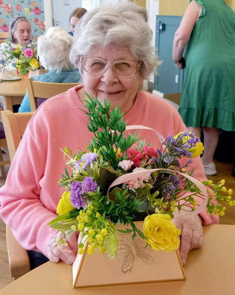 An elderly woman wearing glasses and a pink jumper sits at a table in Lear House, smiling as she holds a floral arrangement in a triangular beige container, displaying her creativity.