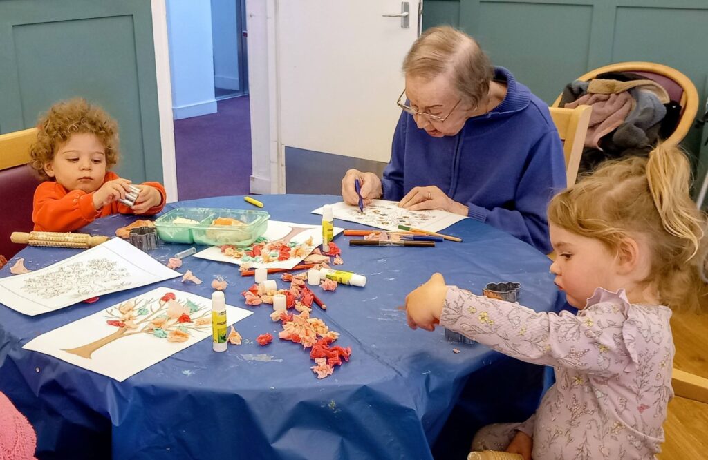 An elderly person and two young visitors sit at a round table, enjoying a wonderful, colourful morning of arts and crafts with paper, glue sticks, and vibrant materials.