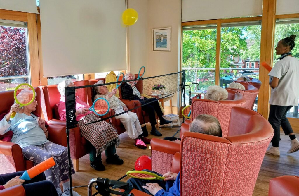 Elderly people seated in armchairs enjoy a lively game of Balloon Tennis across a net indoors, whilst a staff member stands nearby, encouraging flexibility and laughter.