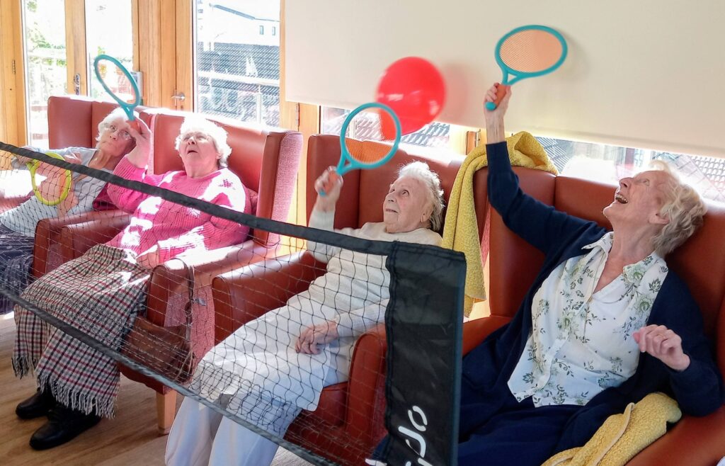 Four elderly women seated in chairs enjoy a game of Balloon Tennis with racquets, divided by a net indoors, encouraging fun and flexibility.
