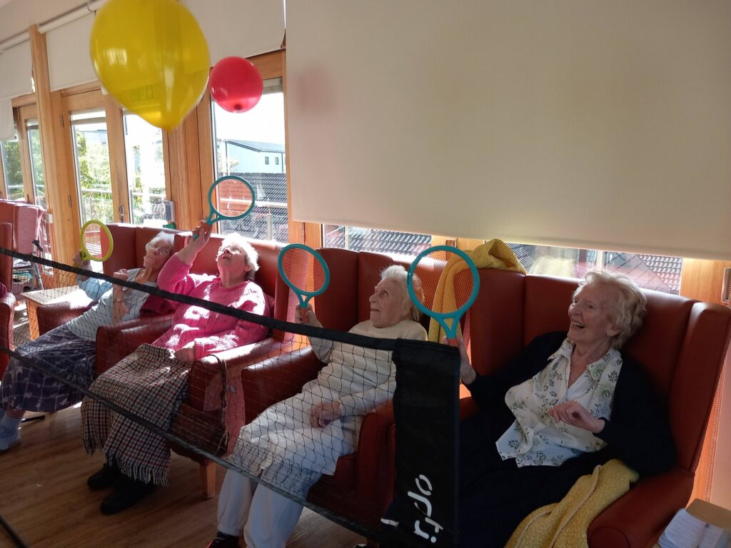 Four elderly women have a lively game of balloon tennis indoors, each holding a racquet and playing over a net, as sunlight pours through large windows.