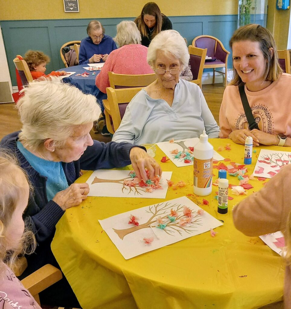 A group of elderly women and young visitors sit at a table making wonderful tree crafts with coloured tissue paper, glue, and felt-tip pens during a colourful morning in a communal room.