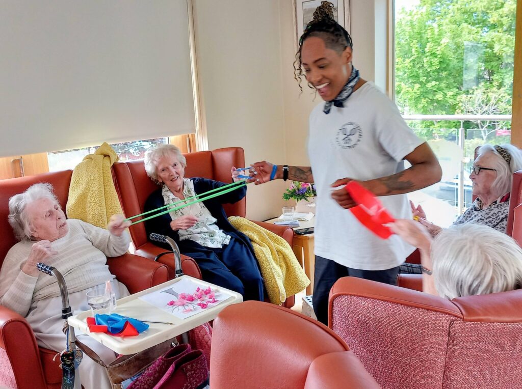 Four elderly women sit in armchairs, enjoying cheerful flexibility exercises with colourful bands, as a staff member leads the session in a brightly lit communal room.