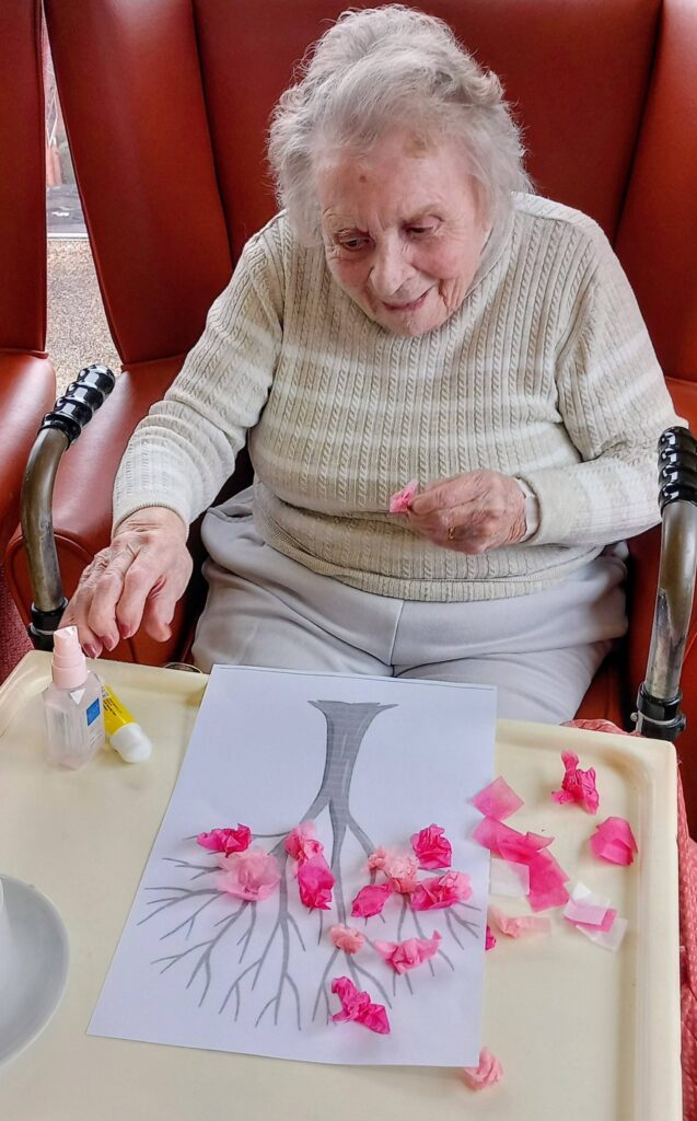 On a Colourful Morning, an elderly woman in a wheelchair creates lovely art by sticking pink tissue paper onto a tree outline on the tray in front of her.
