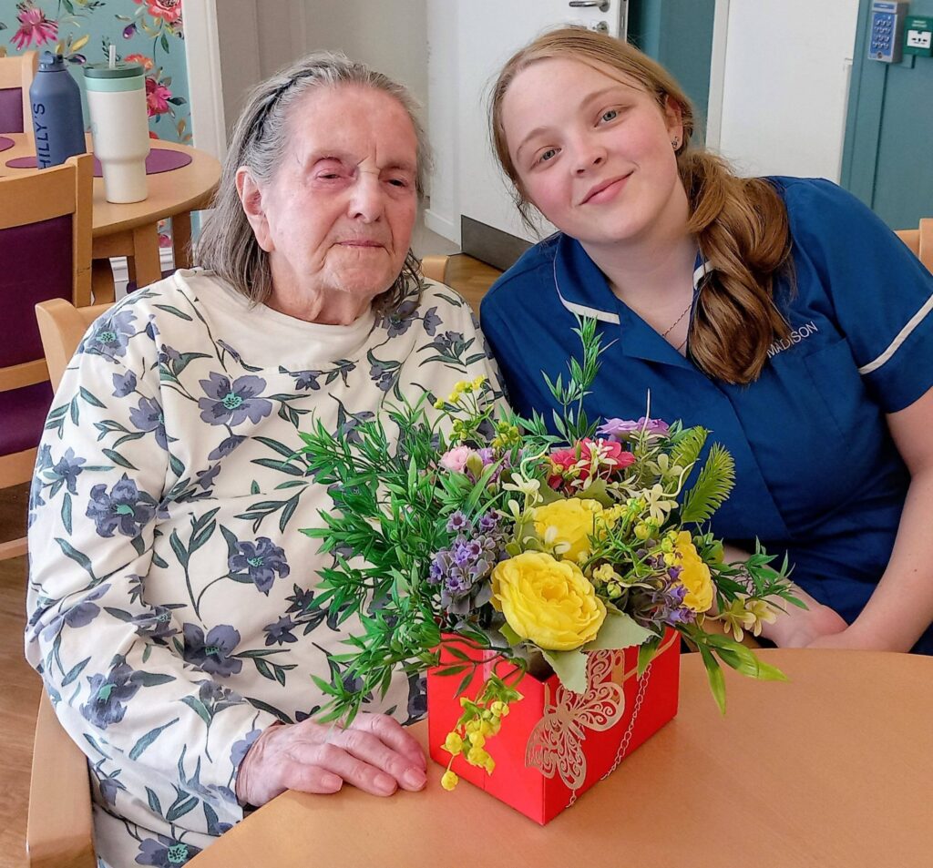 An elderly woman and a young carer sit together at Lear House, their creativity shining as they admire a colourful bouquet of flowers in a red box on the table in front of them.