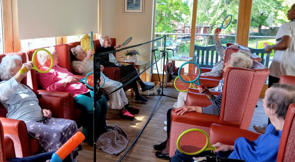 A group of elderly people seated in armchairs play a lively game of indoor balloon tennis with plastic racquets and a net in a well-lit room, encouraging flexibility and enjoyment.