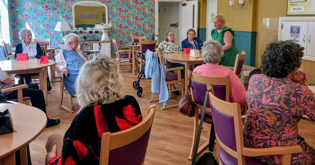 A group of elderly people at Lear House sit round tables in a brightly decorated room, listening to a woman standing at the front who is encouraging creativity and engagement.