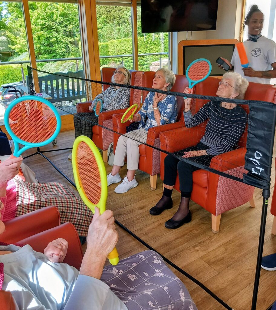 A group of elderly women seated in armchairs enjoy a lively game of balloon tennis with large plastic rackets and an indoor net, assisted by a carer.