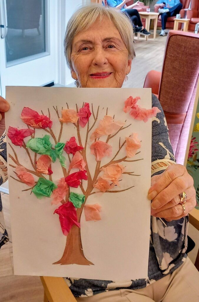 An elderly woman sits smiling on a colourful morning, holding up a drawing of a tree adorned with tissue paper blossoms in various colours.