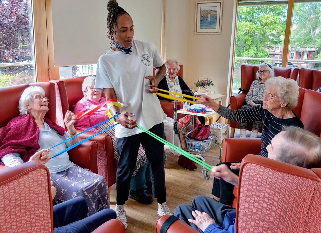 An activity leader guides a group of elderly people seated in chairs as they stretch coloured resistance bands, encouraging fun and flexibility in a bright, cheerful room.